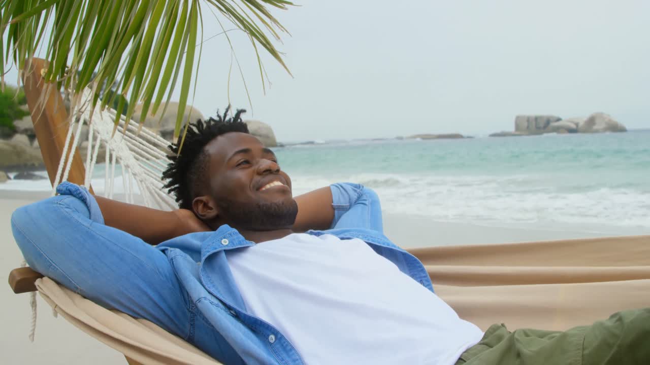 Front view of African american man relaxing in a hammock on the beach 4k