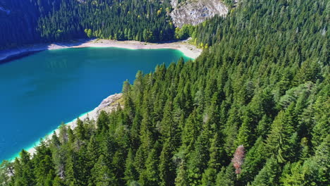 Beautiful-emerald-lake-in-the-middle-of-forest-and-mountains-in-the-background,-aerial-pan-shot