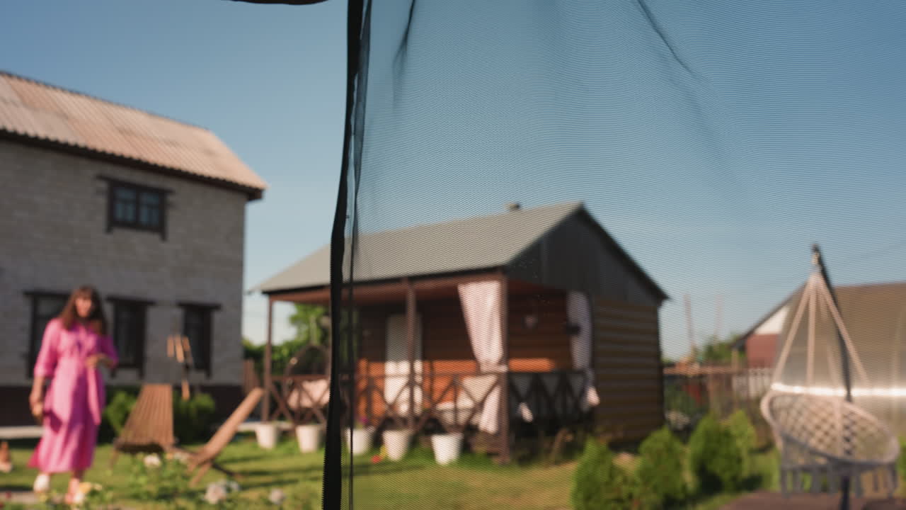 Sunlit Yard Captures Woman In Pink Robe, Woman Strolling Through Yard With Cottage Background In Sunlight, Casual Moment With Woman In Pink Robe Walking Past Cottage Under Warm Sunlight