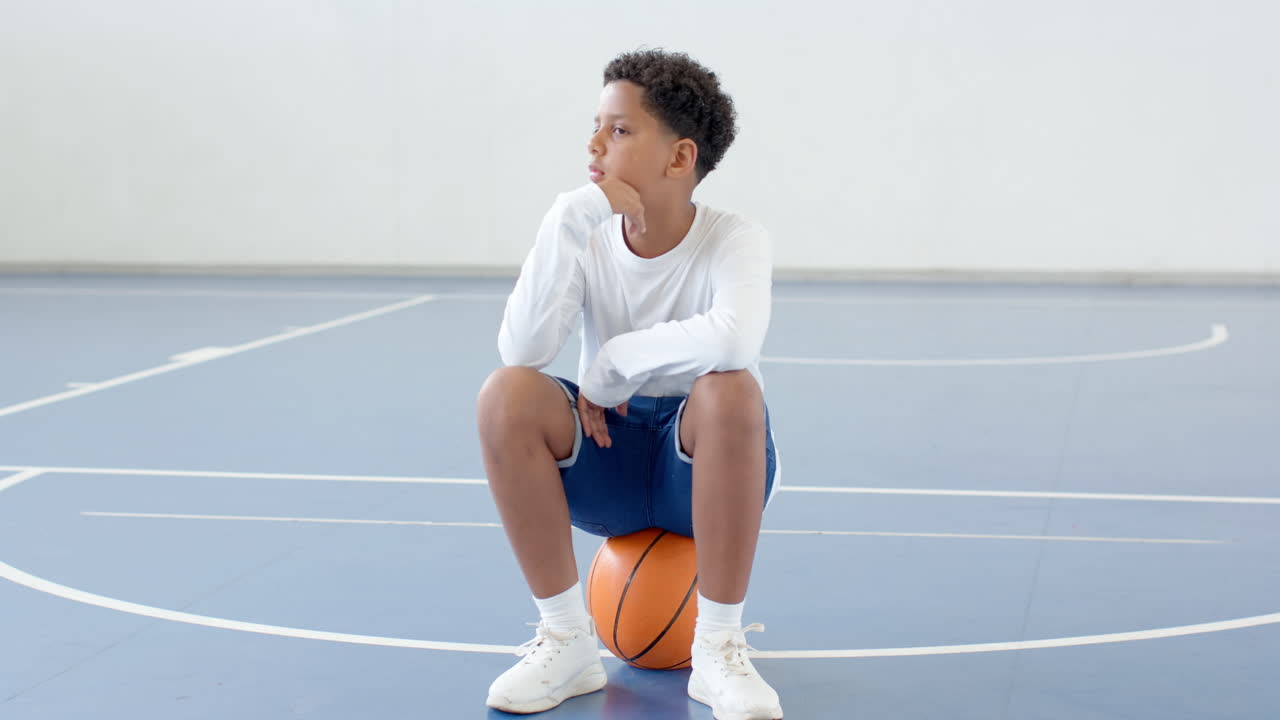 Pensive boy sitting on basketball in gym, contemplating during practice, copy space