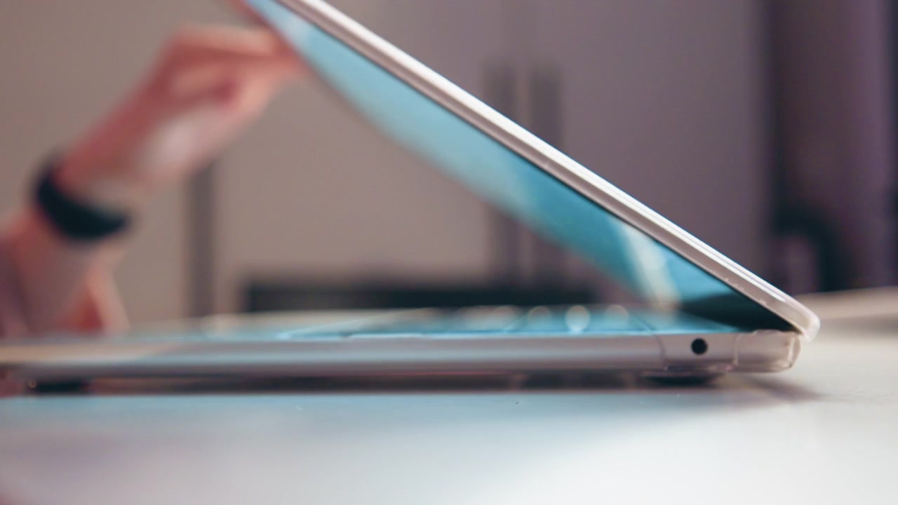 Low-angle close-up of slim laptop resting on light desk, with screen mostly closed, hinge and side port visible, while blurred hand in background suggests reaching, opening, and everyday workspace use