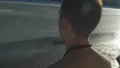 Sunlight shines and reflects on frozen icy surface of remote mountain lake in winter with man, no shirt, sitting outside meditating and preparing to enter frigid water