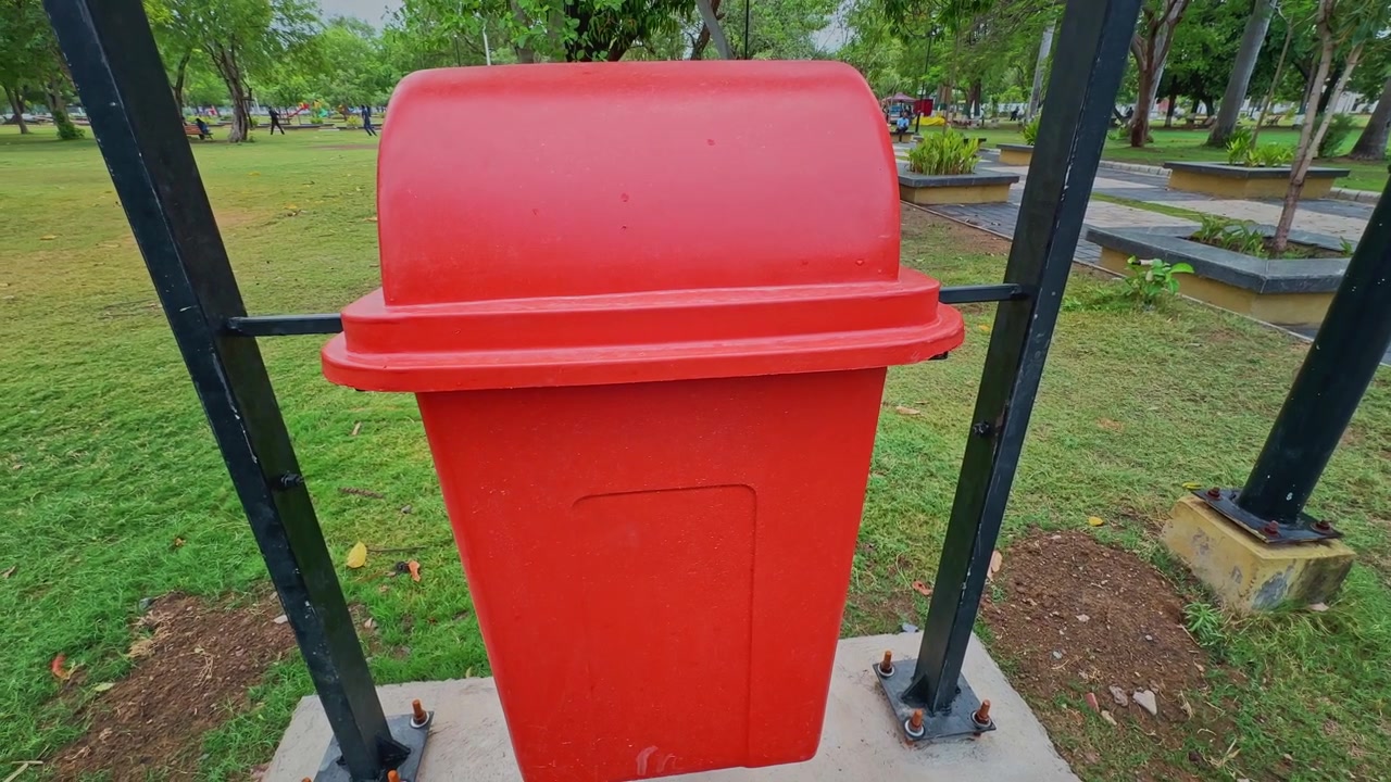 Toma de cámara orbital alrededor de una instalación de cubo de basura rojo brillante en un parque público ajardinado, destacando la infraestructura de limpieza urbana y el entorno recreativo verde.
