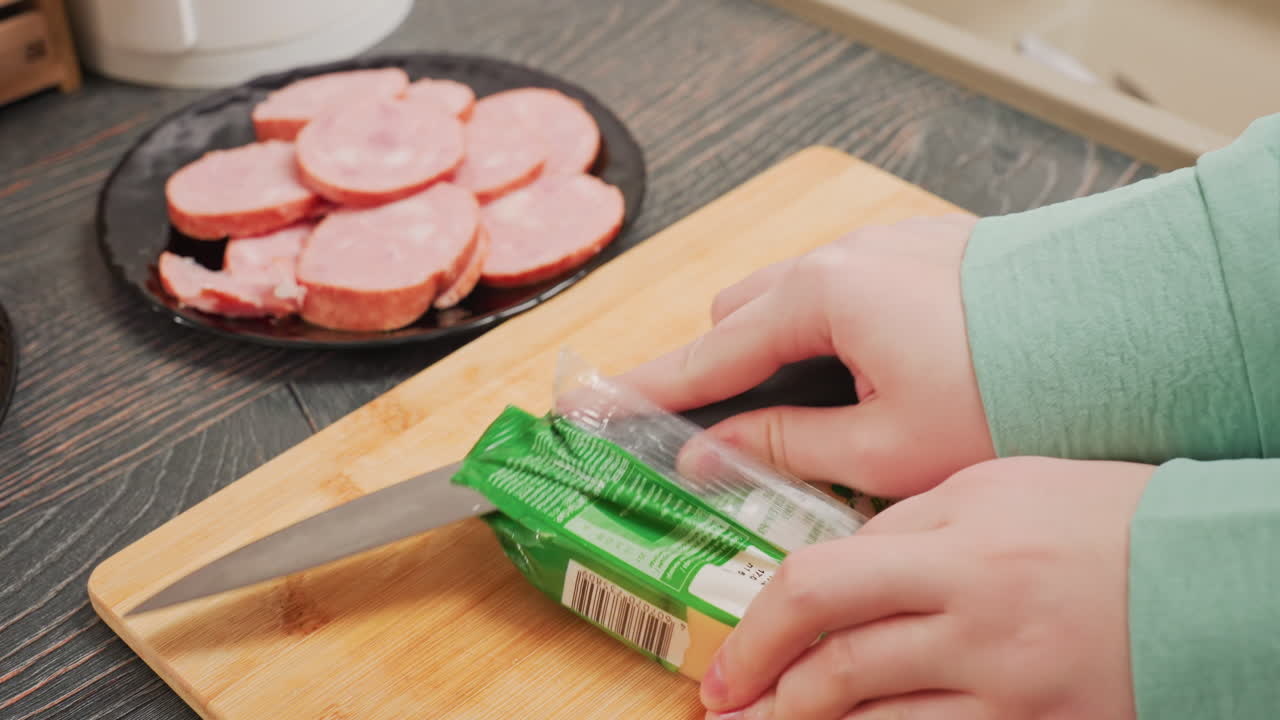 close up of woman in green sleeve trying to open noodles pack with both hands on wooden cutting board before picking up knife to cut it open, with sliced sausage visible on black plate
