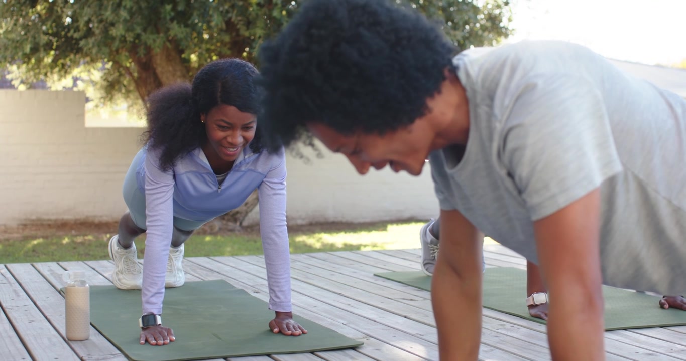 African pair wearing sportswear holding plank on green mats on deck, man cueing, child watching