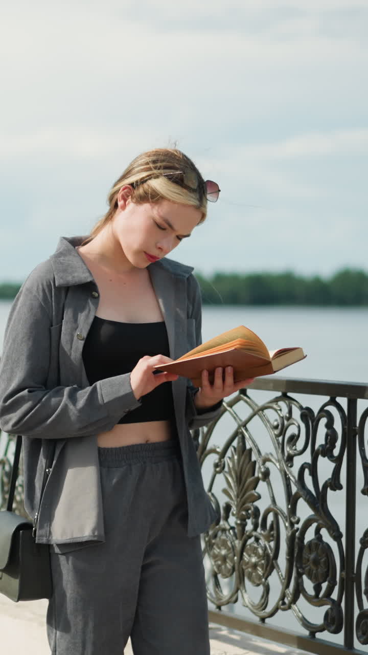 joven con ropa gris y bolso negro de pie cerca de una valla leyendo un libro con enfoque como el viento sopla las páginas y su cabello, los árboles son visibles en la distancia y una orilla del río