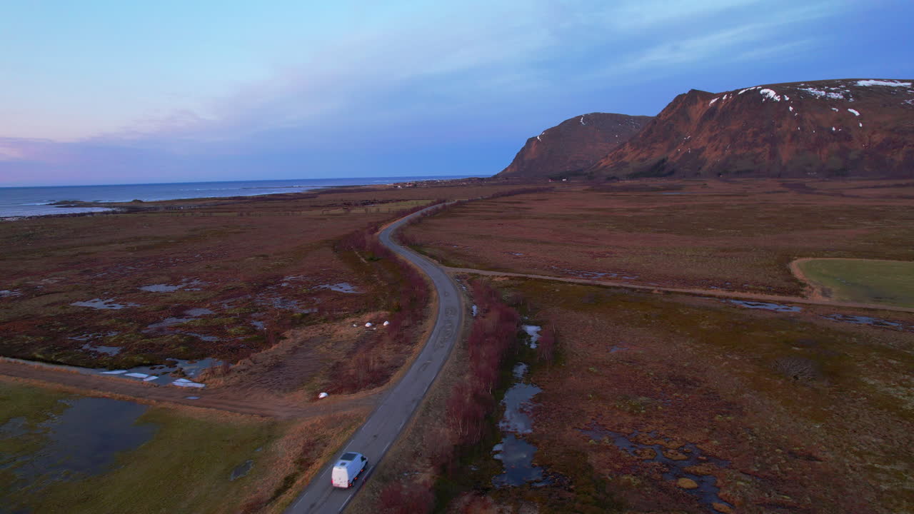 Aerial following view of a van on a road along the coast with mountains and light snow