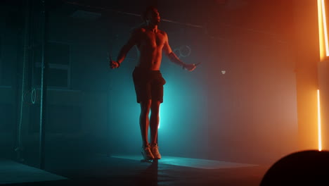Muscular-man-with-bare-torso-is-jumping-using-a-skipping-rope.-Male-boxer-with-skipping-rope.-Fighter-doing-jump-rope-exercises-in-colorful-light-with-smoke-in-slow-motion.