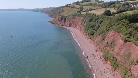 Aerial-flyover-clear-atlantic-ocean-with-rocky-beach,-clay-cliff-and-agricultural-farm-fields-on-top-of-mountain