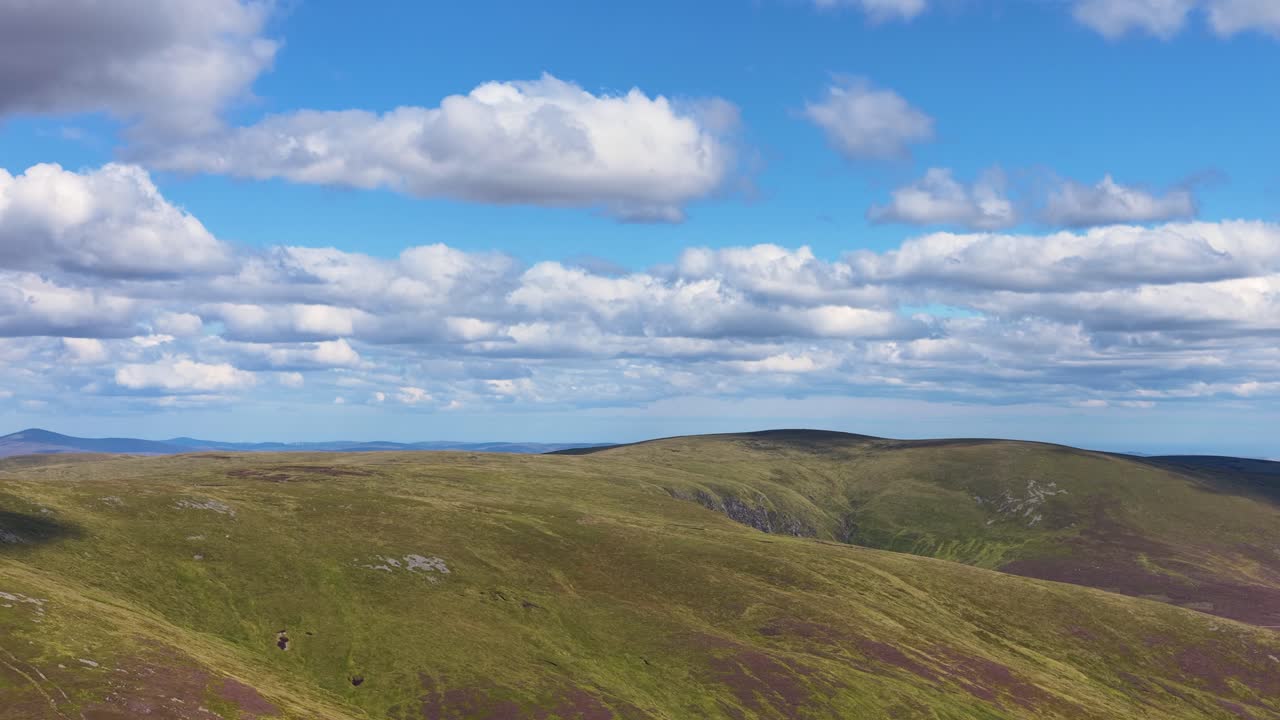 Drone camera glides above green and purple heather hills, bright daylight, tranquil, wide landscape