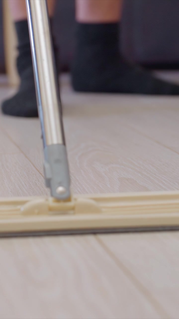 Shows person wearing black socks using wet mop to clean light colored wooden floors. continuous movement of flat mop head across floor grain texture is visible with lower legs and feet