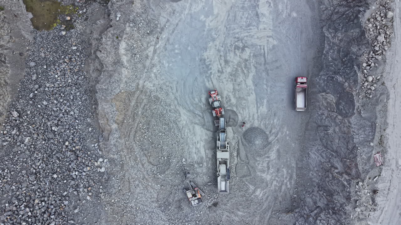 Heavy machinery works in a gravel pit, moving large rocks and materials during the day, with several trucks lined up for transport in an active quarry site