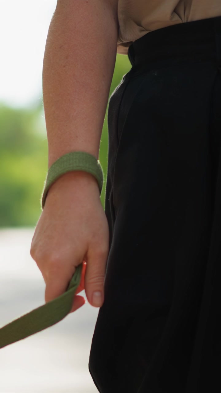 A pessoa carrega um item na mão direita durante um passeio ao ar livre. Uma guia verde é visível em sua mão, com outra por perto. A câmera acompanha a parte inferior do corpo através de um movimento constante para a frente.