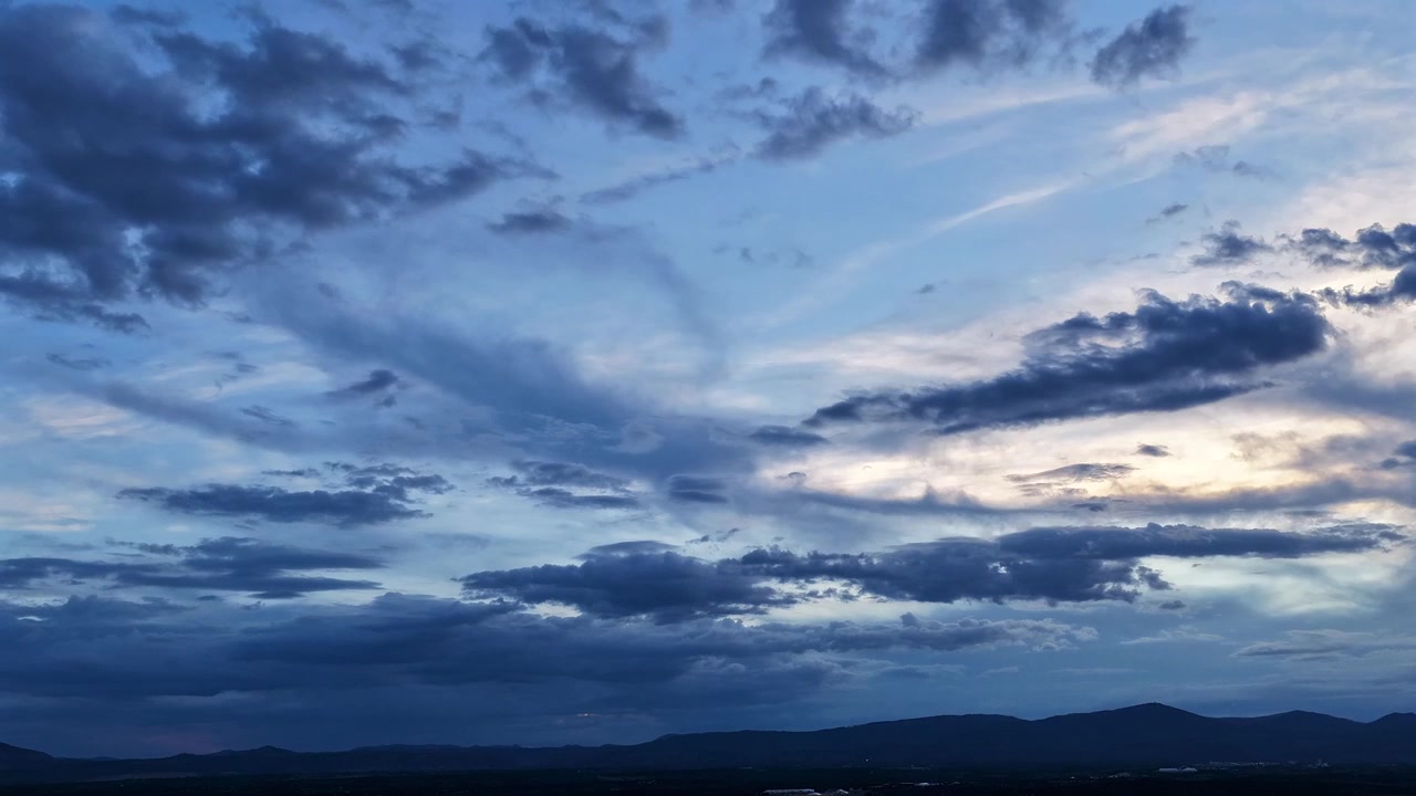4K drone timelapse of dark, moody clouds shifting during blue hour. A cool evening light illuminates the dramatic sky as it drifts over a vast, silhouetted Southwest mountain landscape.