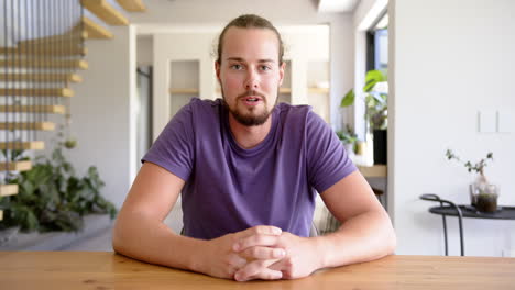 a young caucasian male sits at a wooden table, hands clasped, talking on a video call