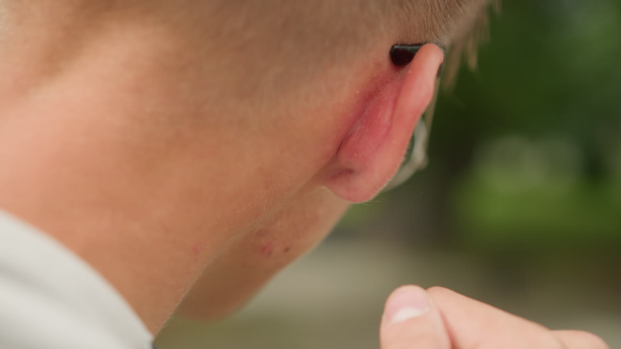 primer plano lateral de la oreja de un músico ajustando un monitor, hombre caucásico con gafas que se ajusta el auricular con los dedos, ambiente de ensayo al aire libre con fondo verde y expresión concentrada