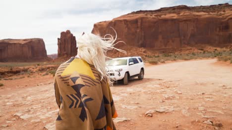 young woman tourist wrapped in brown orange blanket with traditional pattern with blond hair, steps along path at monument valley landscape