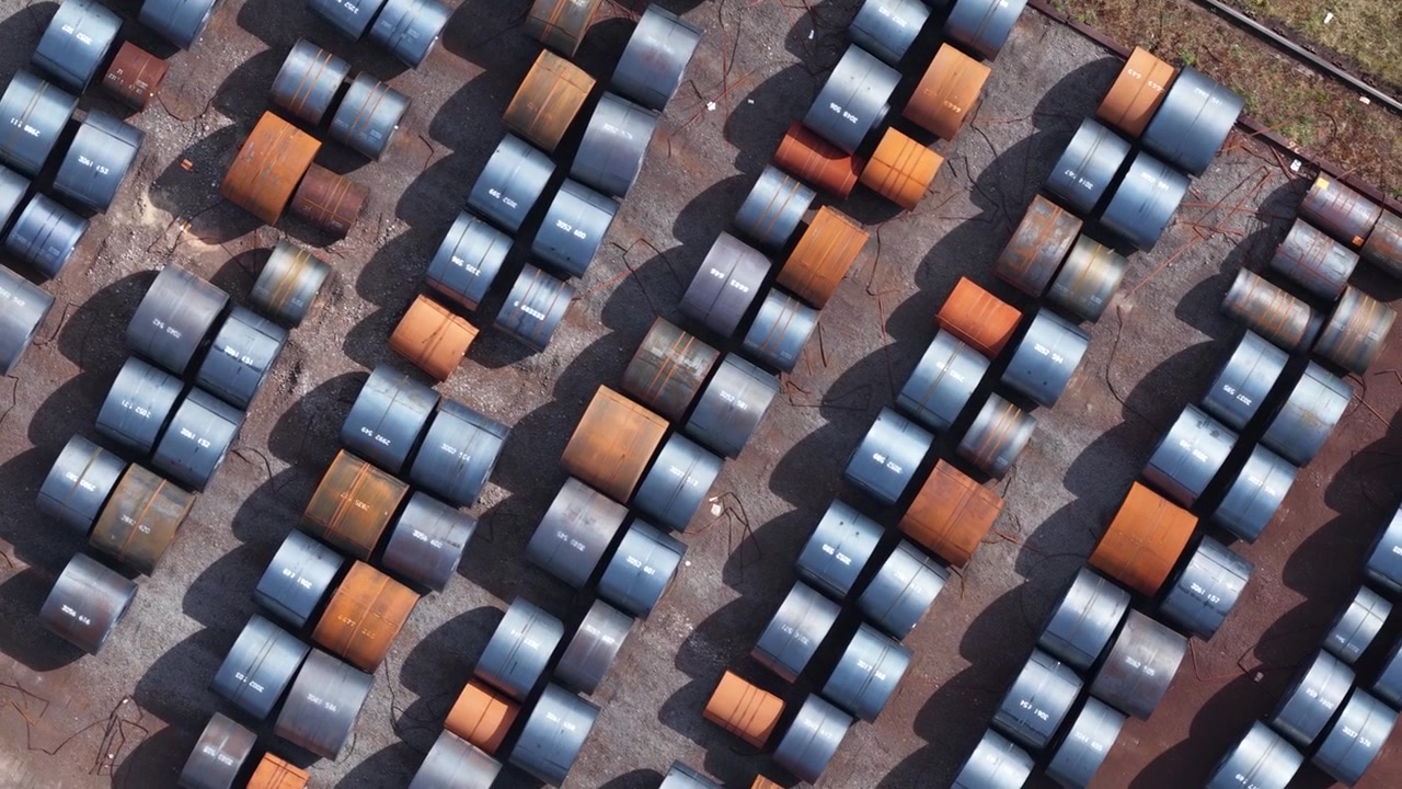 Aerial drone shot capturing rows of steel coils. Including galvanized and rusty ones. Stored in an open air warehouse. Showcasing the industrial scale of metal manufacturing and raw material storage
