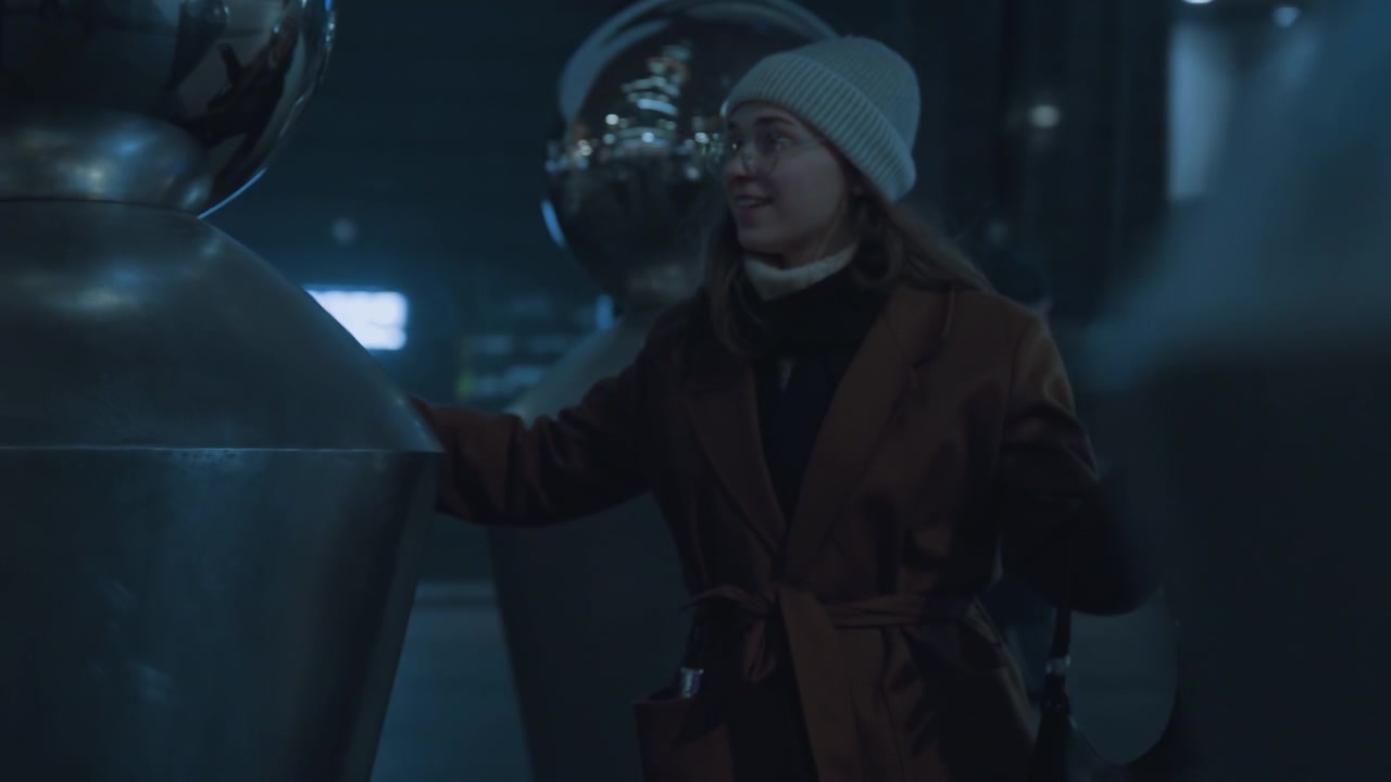 Reflective metal sculpture white woman exploring, touching polished spheres, seeing distorted city reflections, smiling, interacting with public art installation, blue lighting, quiet plaza