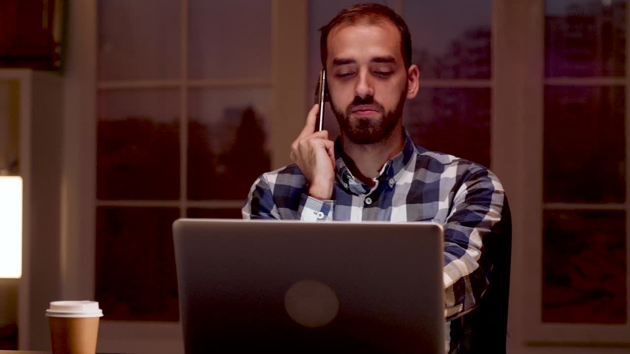 Tired man yawning while on the phone with a laptop in front of him