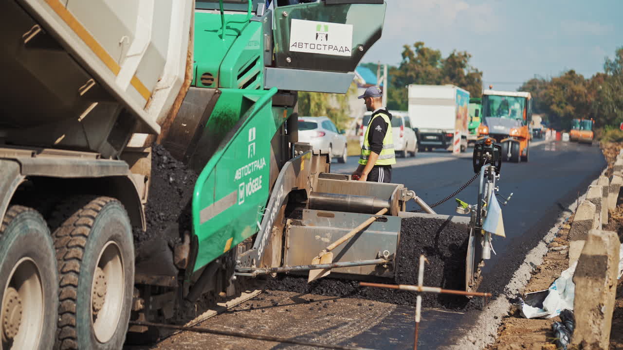 Big green paver machine pouring out asphalt and a worker on the road. Asphalt spreader at work in summer. Male worker with measuring tape during roadworks.