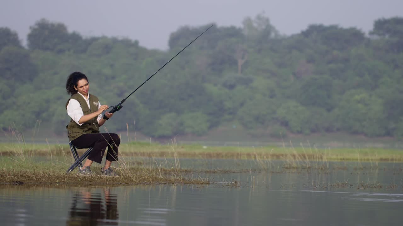 Seorang wanita Asia Selatan memancing di tepi danau yang tenang sambil duduk di kursi, mengangkat tangan, menyesuaikan gulungan pancing, dan melempar joran di siang hari pedesaan yang damai