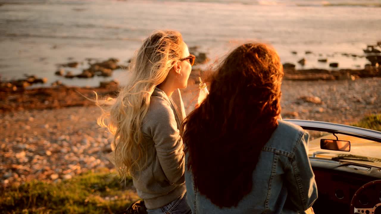 Young women in vintage convertible car drinking soda at beach