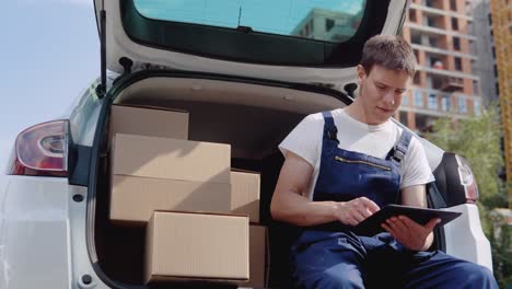 the courier sits on the edge of the open trunk, filled with boxes of goods and fills out documents