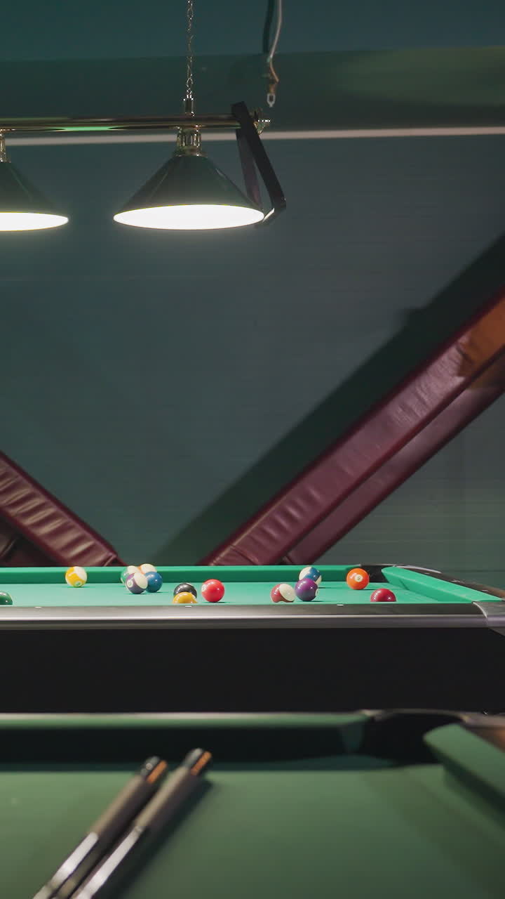 el aprendiz de billar con camisa blanca y pantalones grises se inclina sobre la mesa de billar verde, agarrando el palo de la pilla con un enfoque intenso. fondo oscuro, mientras golpea la pelota de la pila. otra mesa de billa es visible