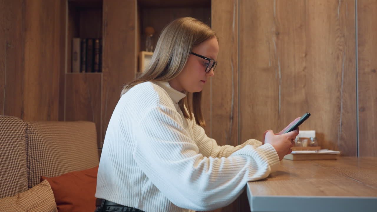 Focused student in white sweater using smartphone while seated at wooden desk in warm cozy room with soft lighting, shelves in background, and study materials nearby