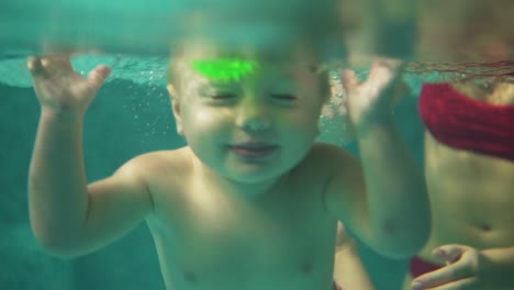 cute blonde toddler is diving under the water in the swimming pool to get his toy while his mother is teaching him how to swim