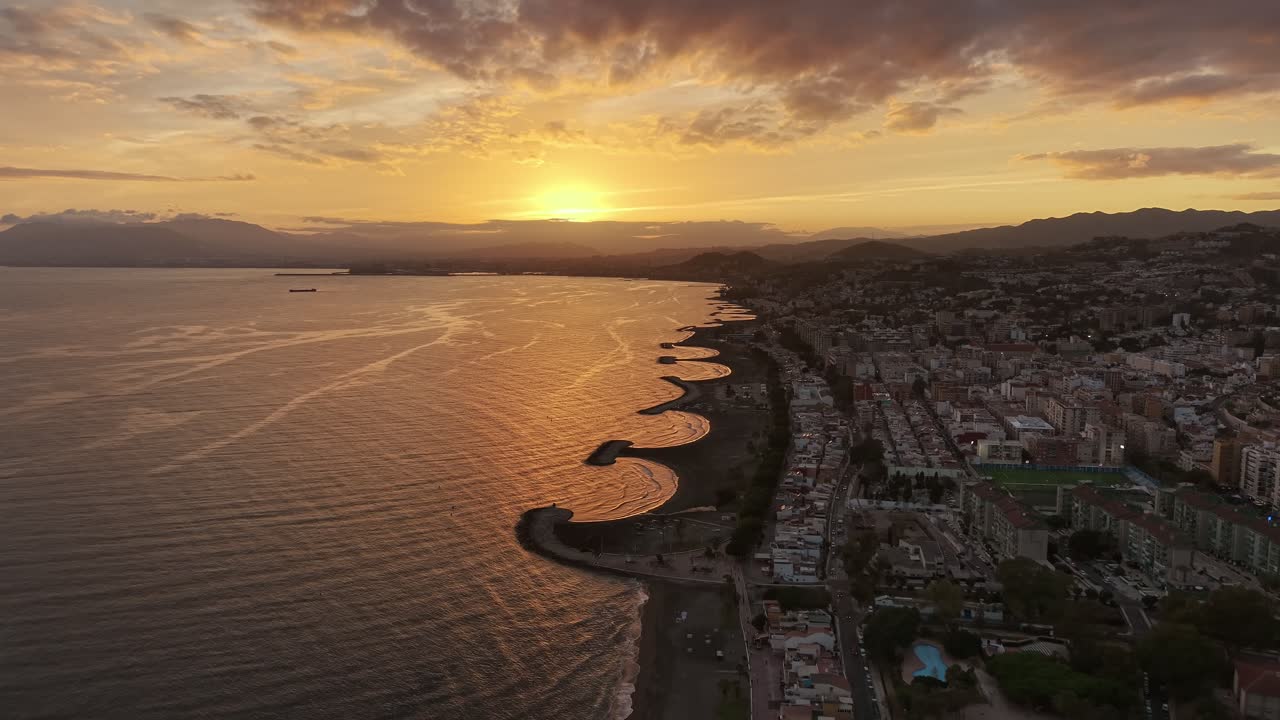 Malaga coastline during sunset showing curved beaches, calm waves, and the city glowing under golden light - Aerial