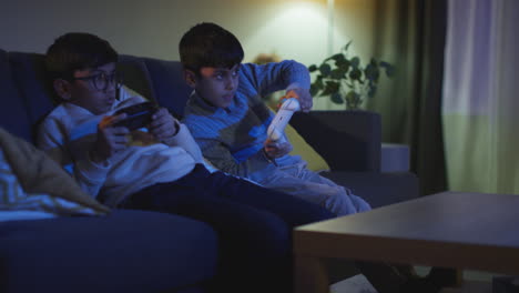 two young boys sitting on sofa at home playing with computer games console on tv holding controllers late at night 3