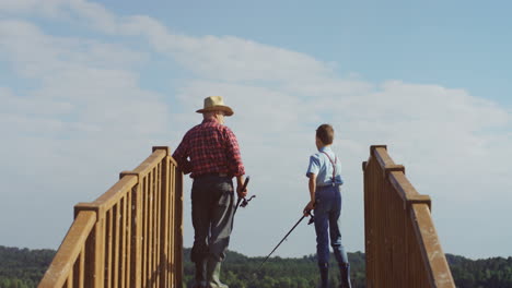 rear view of senior man in a hat standing on the wooden bridge with little boy and fishing with rods in hands