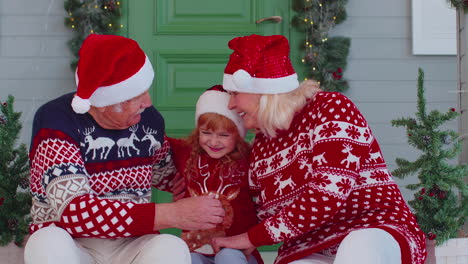 Senior-grandparents-with-granddaughter-in-Santa-Claus-hat-celebrating-Christmas-near-decorated-house