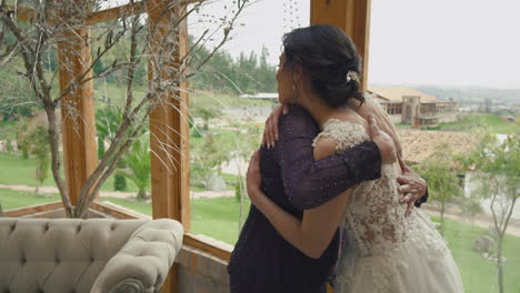 touching clip of a bride tenderly hugging her mother, near a window overlooking a beautiful natural landscape.
