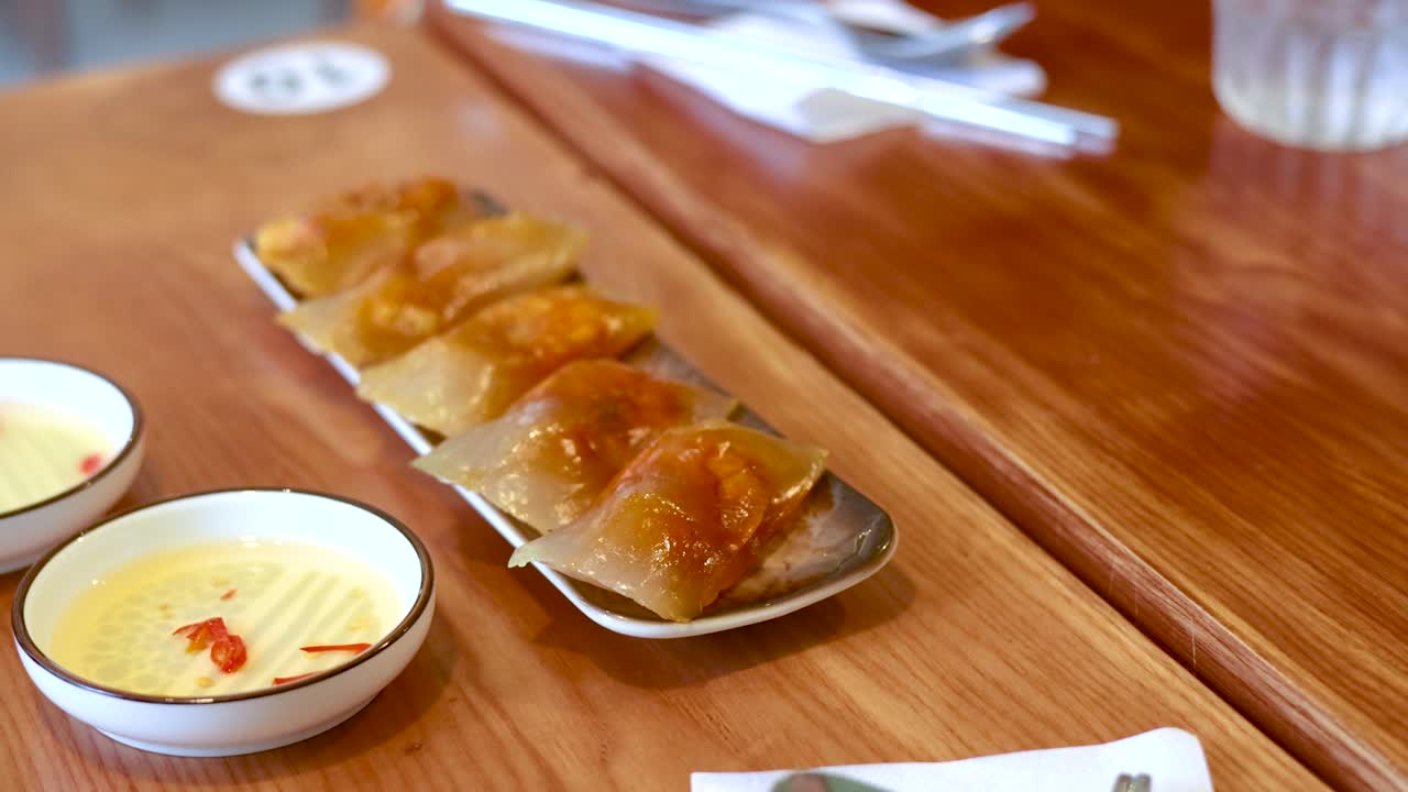 Static high-angle shot of translucent shrimp dumplings and dipping sauce on a wooden restaurant table
