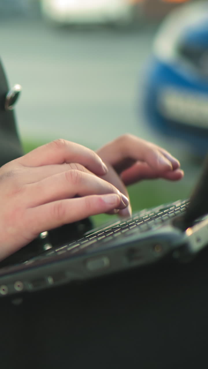 cerca de las manos escribiendo en la computadora portátil al aire libre mientras usa un traje negro con botones plateados, contra un fondo borroso con coches estacionados y vegetación