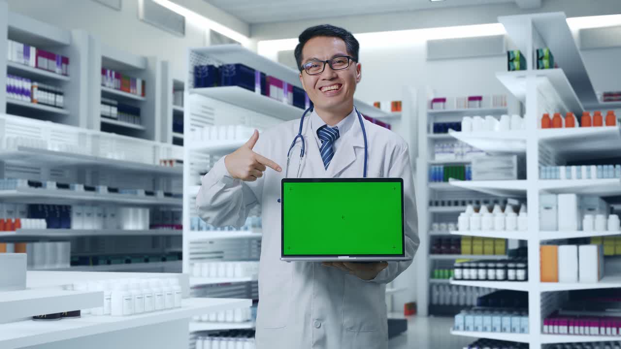 스테토스코프를 들고 있는 아시아 남성 의사 가 약국 에 서 있는 동안 녹색 화면 노트북 을 가리키고 미소 짓고 있다