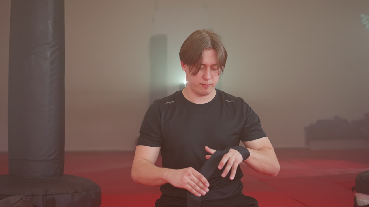 Boxer sitting on red mat in gym wrapping hands with black hand wraps before training, preparing for sparring session with focused expression, punching bags and sports gear around creating intense atmosphere