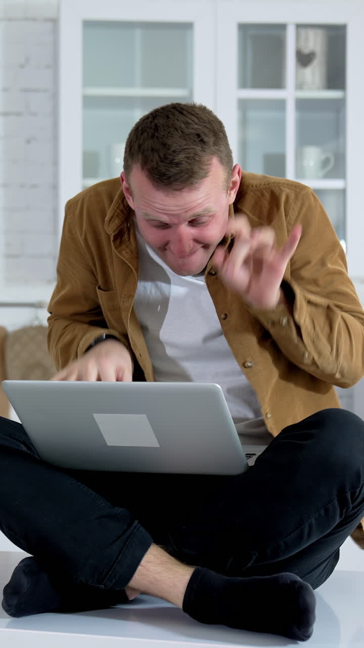 Young man sitting on a kitchen table with a laptop. Nervous freelancer man sitting at table looking at laptop computer screen typing quickly feeling mad. Vertical video