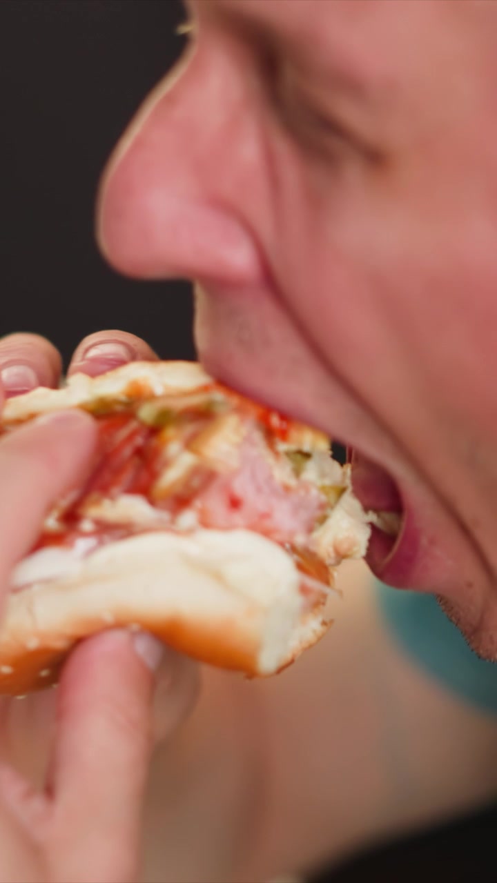 Close up of man eating food focusing on his mouth and hand as man takes bites. subject holds sandwich with one hand while bringing it to his mouth repeatedly. Background is dark emphasizing