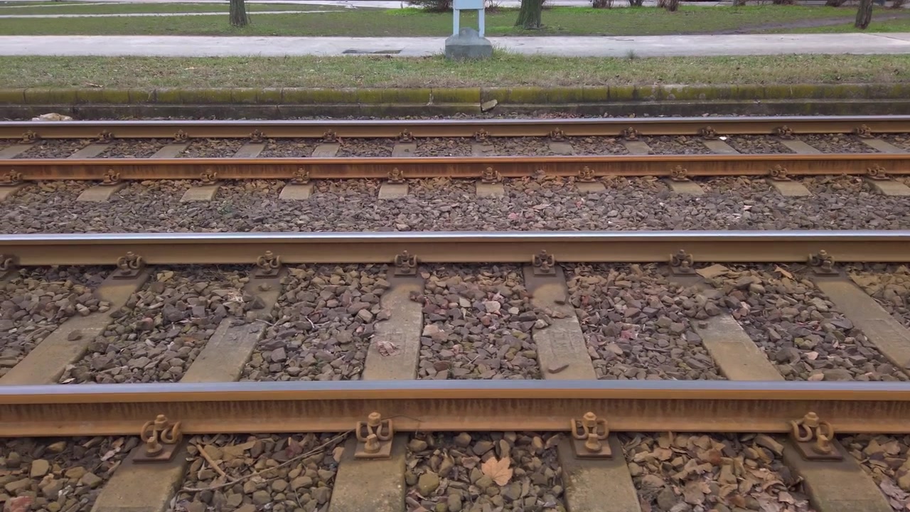 Handheld first person tracking shot moves horizontally along empty parallel steel tram tracks with stone ballast and concrete ties by an urban sidewalk path on a cloudy winter day in Budapest.