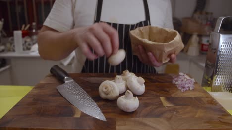 chef puts out white mushrooms onto wooden cut board from paper bag in the kitchen