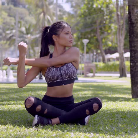 sporty woman sitting with crossed legs and stretching arms in park