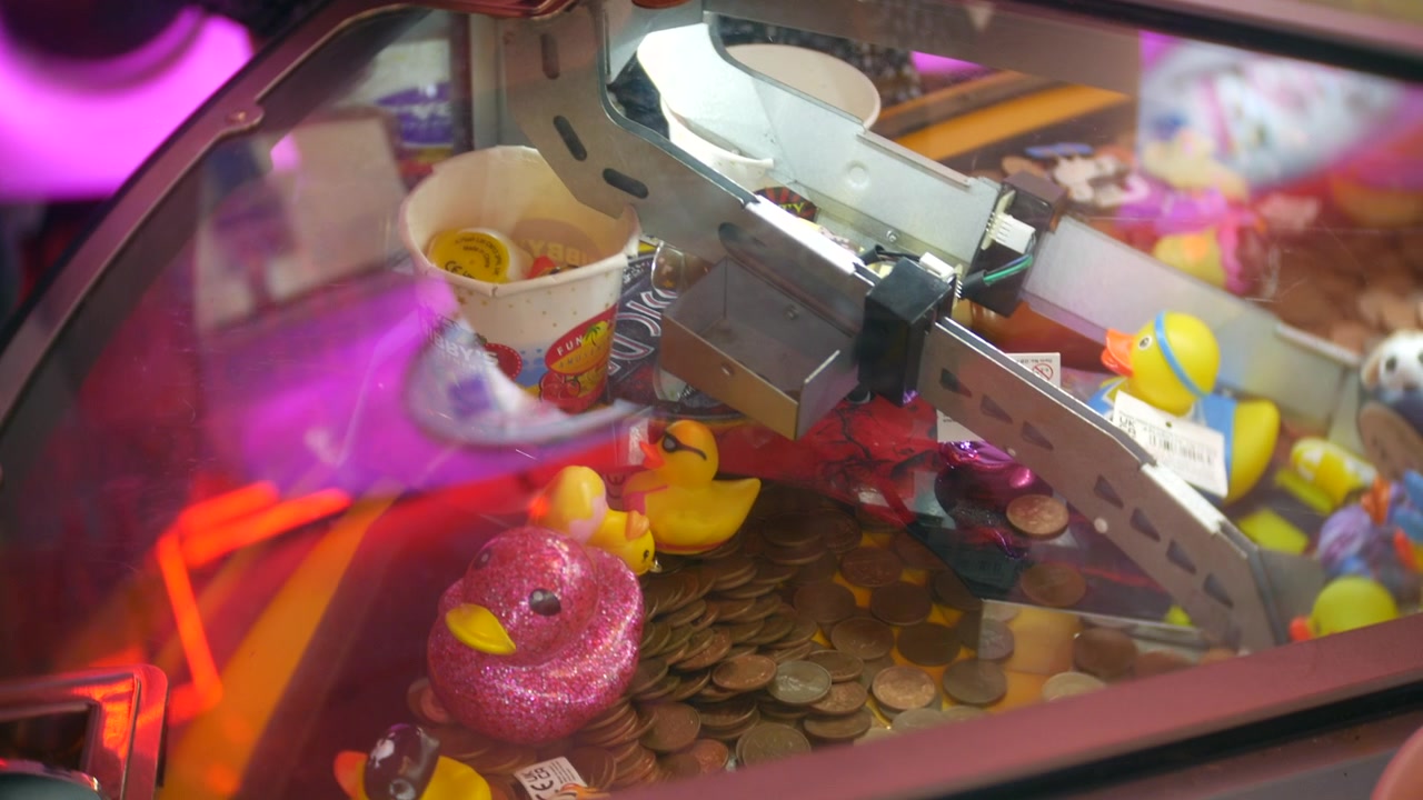 Child operating an arcade claw machine grips the metal control while prizes sit beneath the glass. Close-up view shows hands, bright pink lighting, and colorful toy capsules