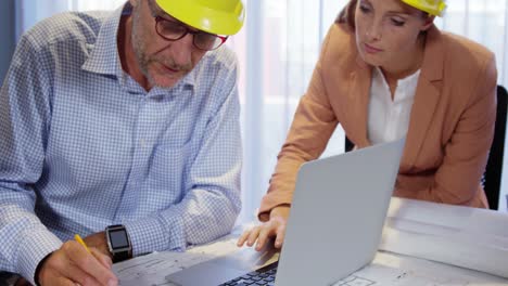 Businesswoman-using-laptop-while-coworker-working-on-blueprint