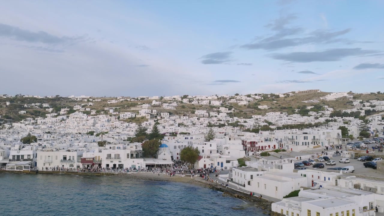 Vista drone dell'affollata spiaggia del porto di Mykonos città, con folla lungo il lungomare, una chiesa dalla cupola blu e case bianche che si arrampicano sulle colline.