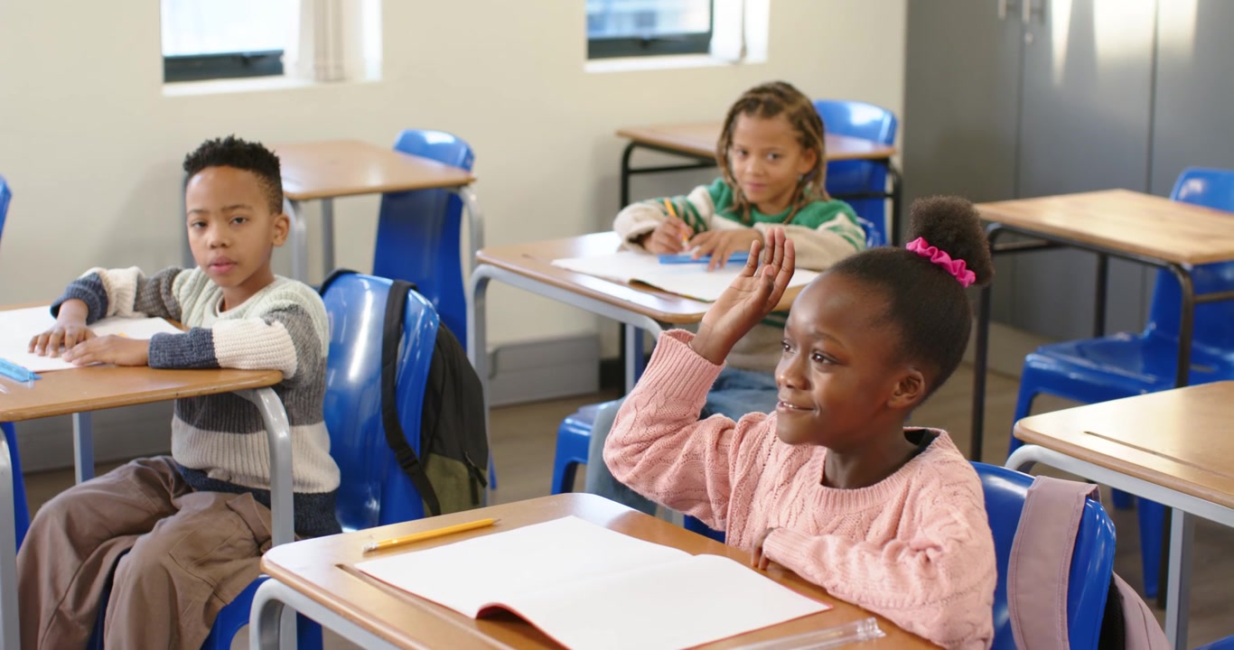 African kids sitting at desks by windows as teacher asking question, girl raising hand
