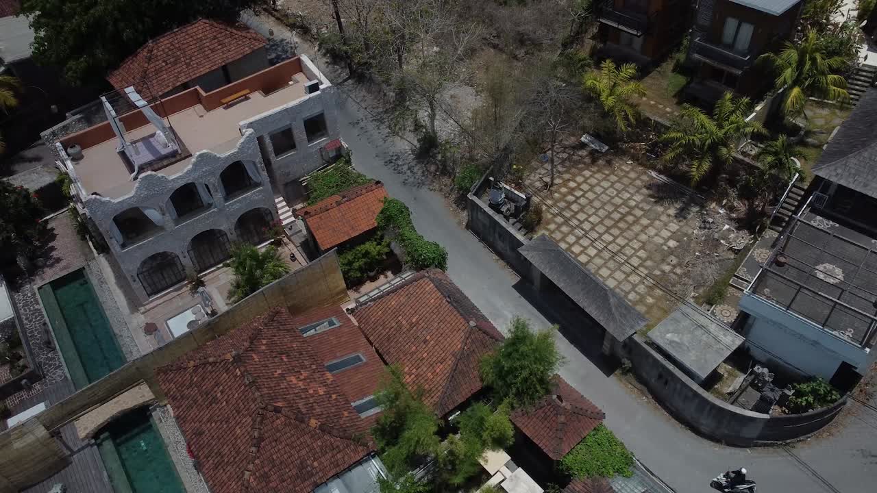 A high-angle 4K drone view captures a dusty, winding back street in Uluwatu, Bali, lined with traditional and modern villas. A lone scooter speeds down the quiet residential road